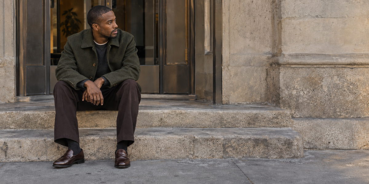 Editorial photograph of a seated model in a dark jacket and trousers outside a stone building